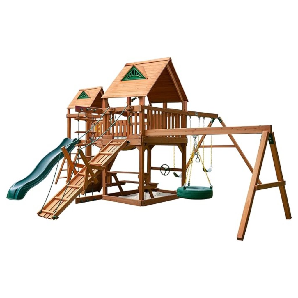 The Gorilla Playsets Pioneer Peak Treehouse Wooden Swing Set includes a tire swing, clatter bridge and tower, two elevated playhouses with green roofs, slide, climbing ramp, two swings, and rope ladder for backyard fun.